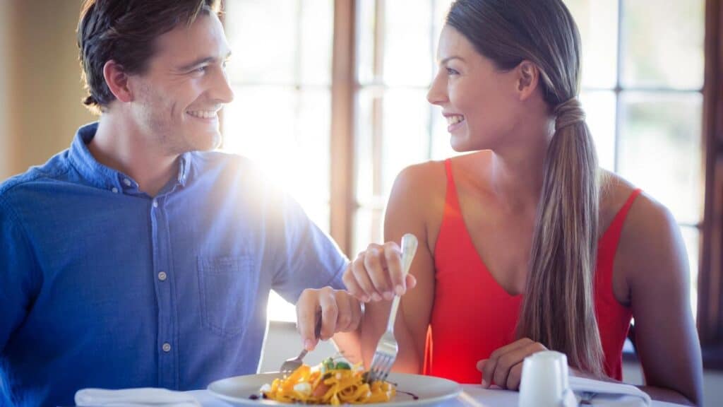 A man and a woman sit at a table in a sunlit room, smiling at each other while sharing a split entree of pasta&mdash;enjoying economy and togetherness with every bite.