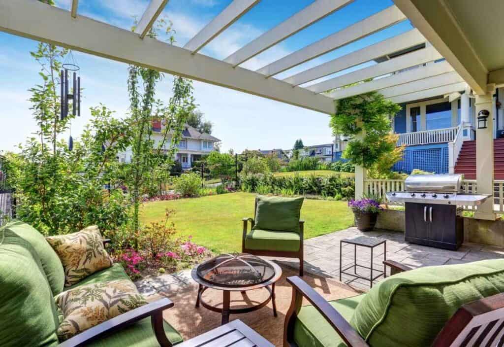 Patio with green cushioned furniture, a fire pit, and a grill under a pergola creates an inviting outdoor living space, overlooking a landscaped backyard and neighboring houses on a sunny day.