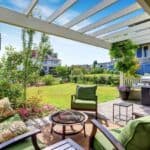 Patio with green cushioned furniture, a fire pit, and a grill under a pergola creates an inviting outdoor living space, overlooking a landscaped backyard and neighboring houses on a sunny day.