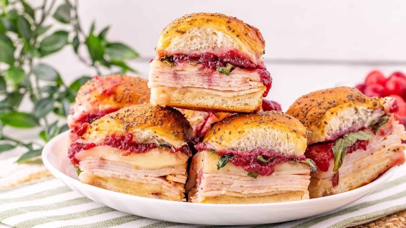 A stack of turkey sliders on buns topped with poppy seeds, filled with turkey, melted cheese, spinach, and cranberry sauce.