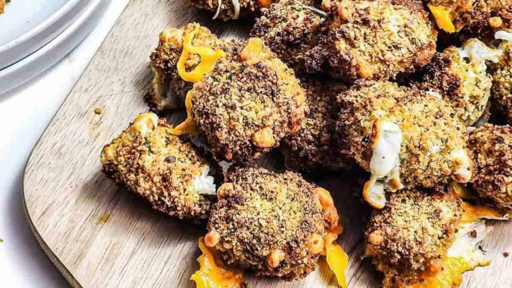 A close-up of several breaded and baked cheese-stuffed jalape&ntilde;o poppers on a wooden cutting board.