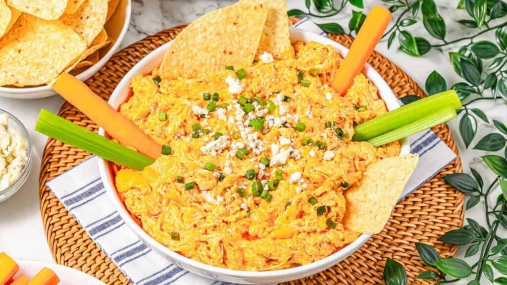 A bowl of buffalo chicken dip topped with chopped green onions and cheese, garnished with celery sticks, carrot sticks, and tortilla chips.
