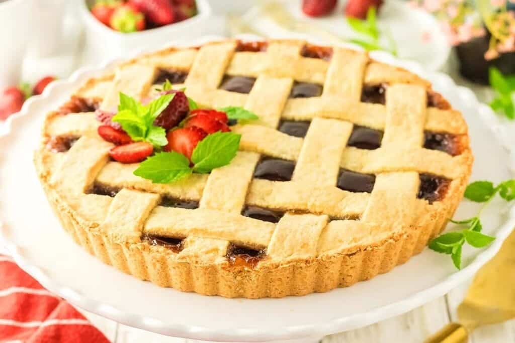 A lattice-topped fruit pie garnished with fresh strawberries and mint leaves on a white cake stand, with strawberries and greenery in the background.