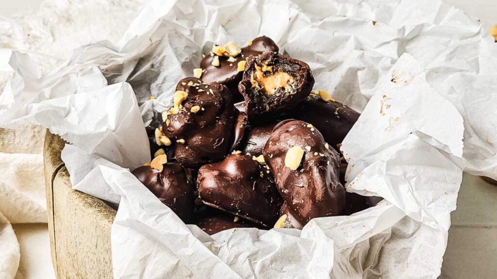 A pile of chocolate-covered treats with a creamy filling and sprinkled nuts, arranged on crumpled parchment paper in a wooden bowl.