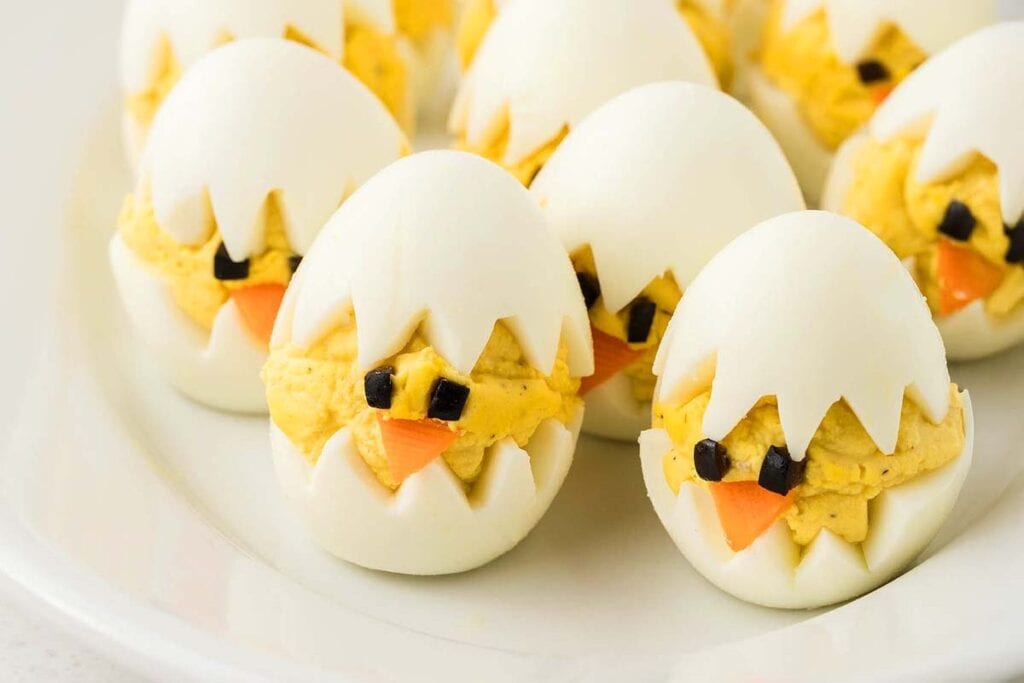 Deviled eggs styled to look like baby chicks hatching, with cracked egg white tops, yolk filling, and faces made from small pieces of olives and carrots.