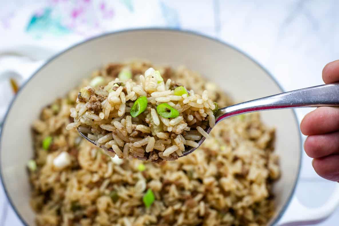 A close-up of a spoon holding dirty fried rice mixed with ground meat and sliced green onions above a pot of the same dish.