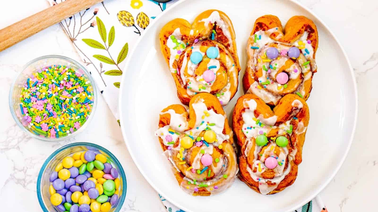 Four Easter bunny-shaped pastries with icing and colorful candies rest on a white plate. Nearby, two bowls of sprinkles and candies, a spatula, and a floral cloth add to the festive scene&mdash;perfect for savoring delightful cinnamon rolls this springtime.