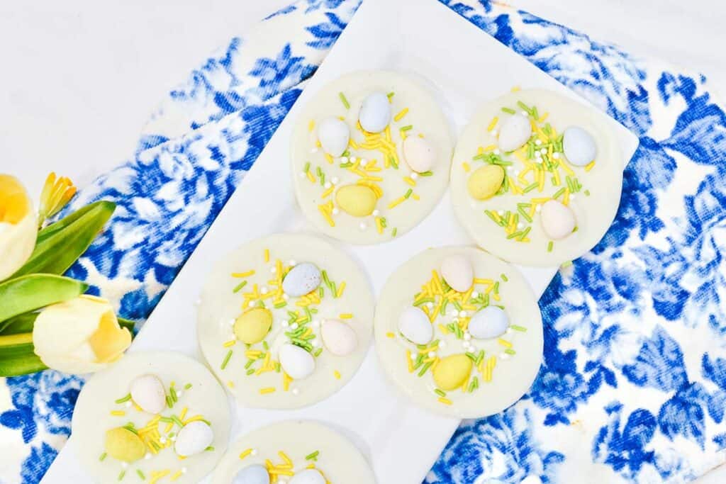 White chocolate discs topped with pastel candy eggs and yellow sprinkles are arranged on a rectangular white plate next to yellow tulips and a blue patterned cloth.