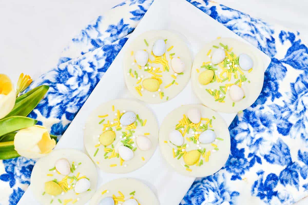 White chocolate disks topped with small pastel eggs and sprinkles sit on a tray. Nearby, there are yellow tulips and a wooden bunny figure on a blue floral cloth.