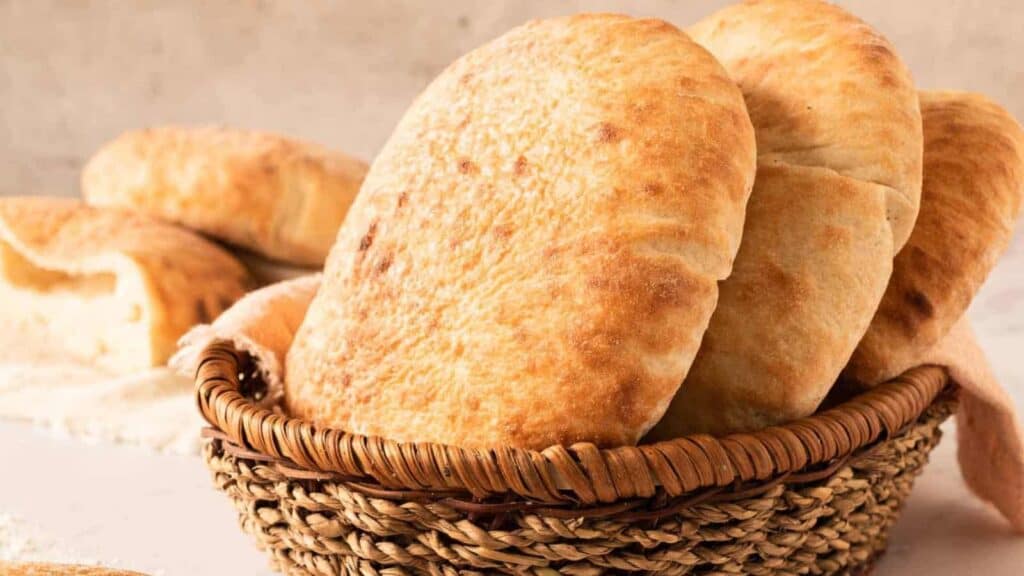 A woven basket filled with several pieces of round, golden-brown flatbread sits on a light surface with more bread in the background.