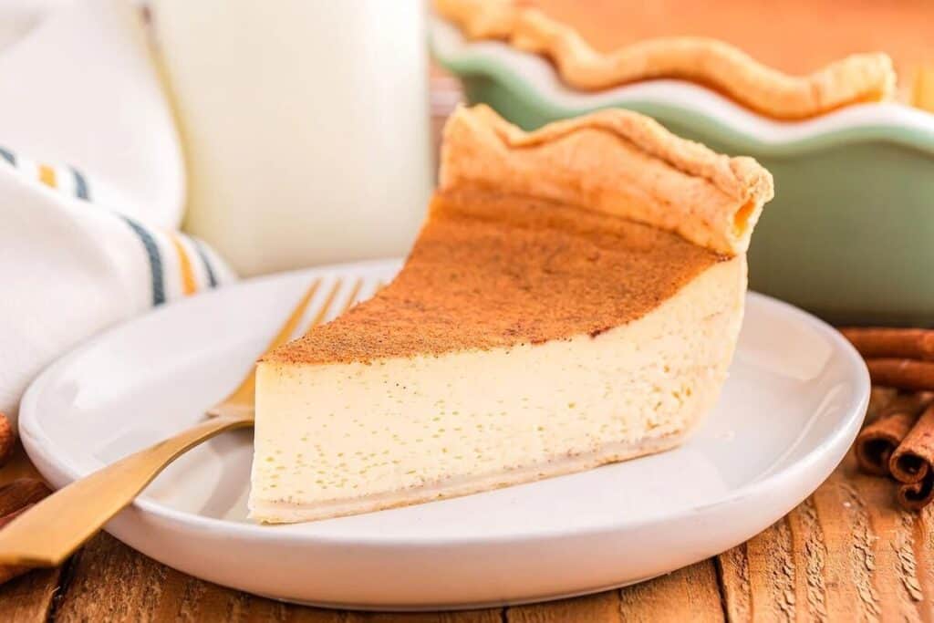 A slice of custard pie with a golden-brown crust and cinnamon topping sits on a white plate with a gold fork.