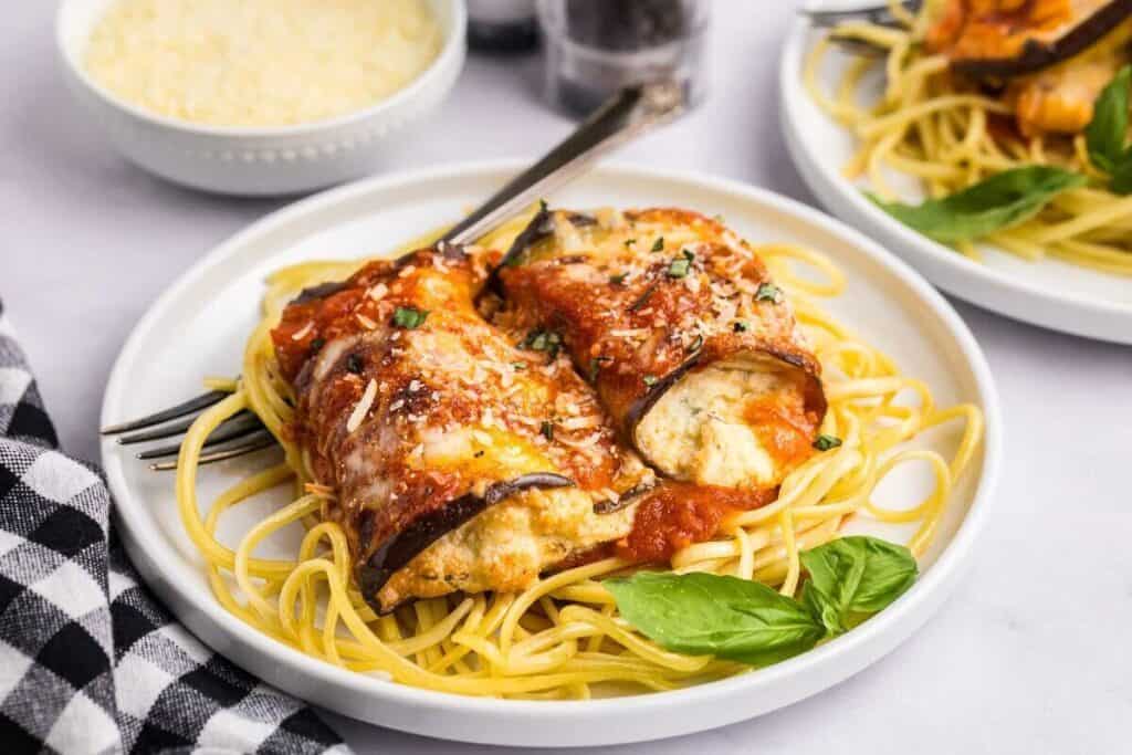 A plate of spaghetti topped with eggplant rollatini, marinara sauce, grated cheese, and fresh basil, with a checkered napkin and a bowl of cheese in the background.
