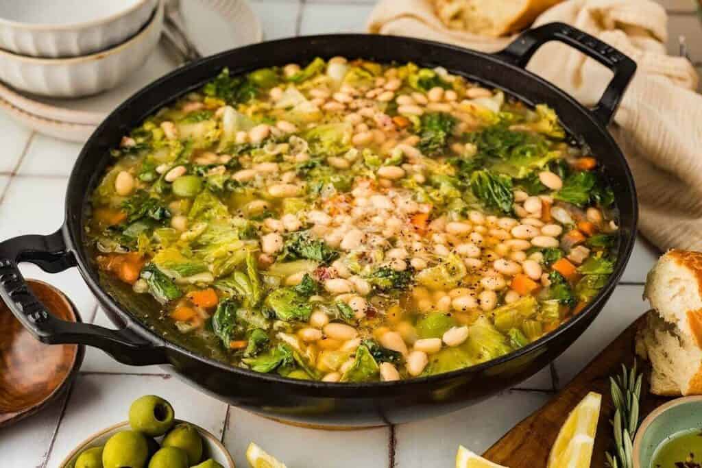 A large black pot filled with vegetable and white bean soup, garnished with grated cheese and black pepper, sits on a table with bread, olives, and lemon wedges nearby.