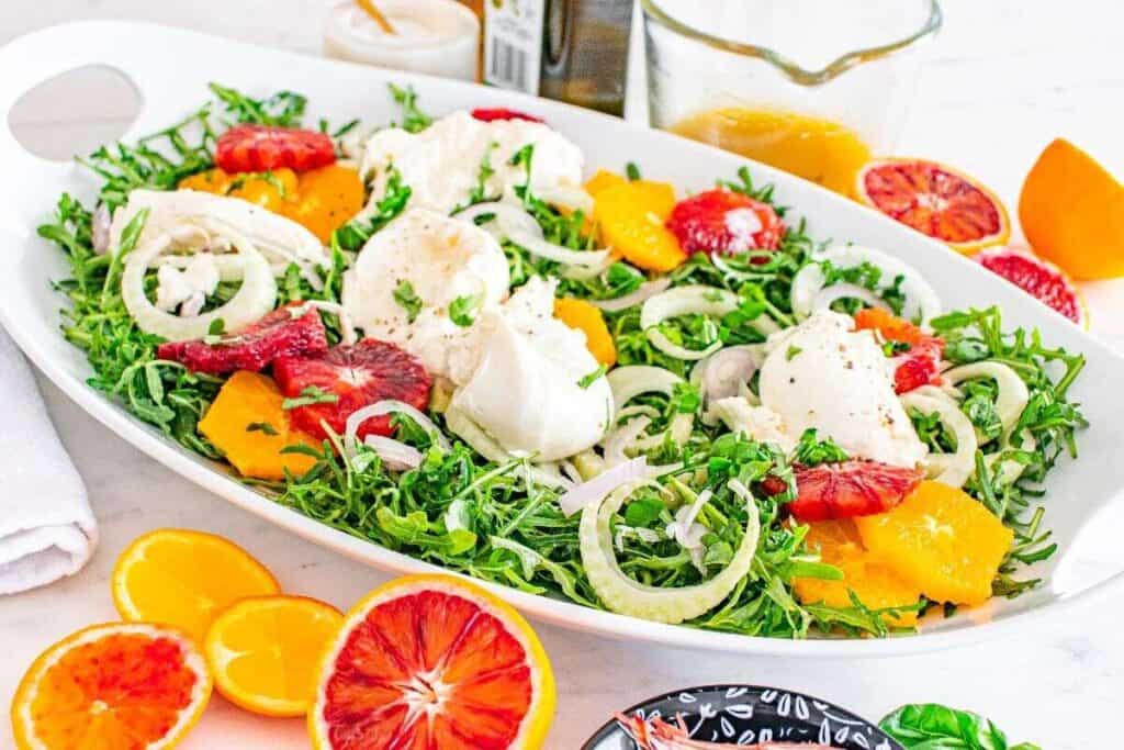 A large white platter with a salad containing leafy greens, sliced citrus fruits, fennel, red onion, and dollops of cheese, with dressing and citrus halves nearby.
