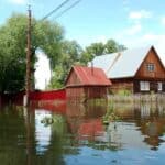 A wooden house and outbuilding are surrounded by floodwater, with partially submerged plants and trees visible under a partly cloudy sky, highlighting spring flood concerns.
