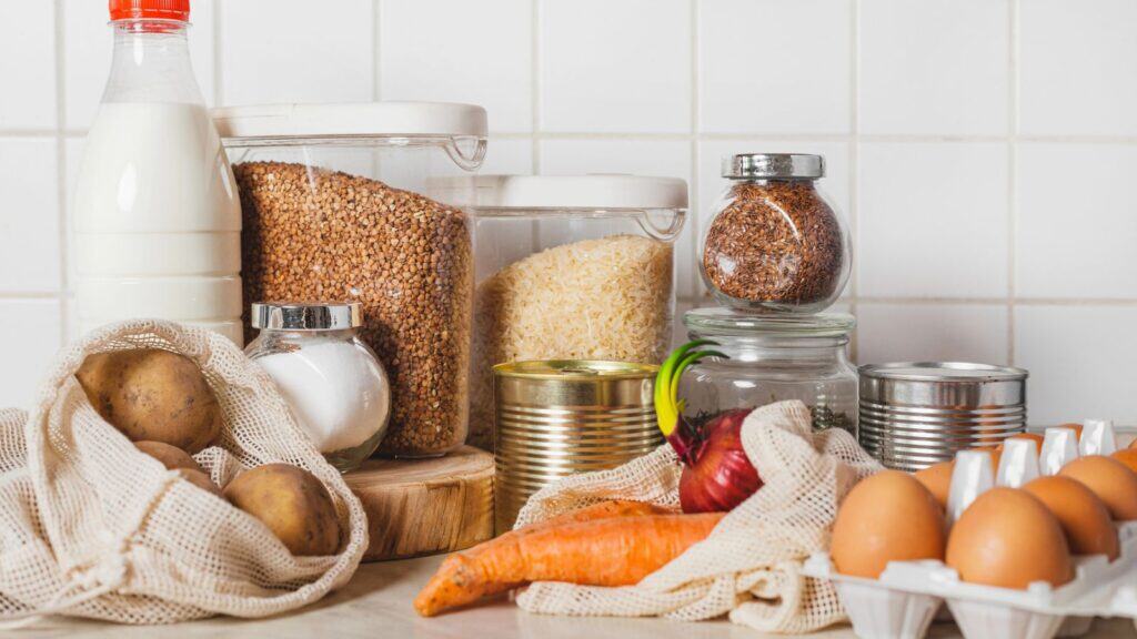 A variety of pantry staples including grains, canned goods, eggs, vegetables, and a bottle of milk are arranged on a kitchen counter against a tiled backsplash.