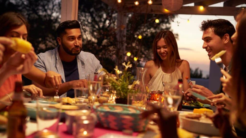 A group of people sit around a table outdoors at dusk, sharing a meal and smiling, with string lights overhead and food and drinks on the table.