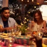 A group of people sit around a table outdoors at dusk, sharing a meal and smiling, with string lights overhead and food and drinks on the table.