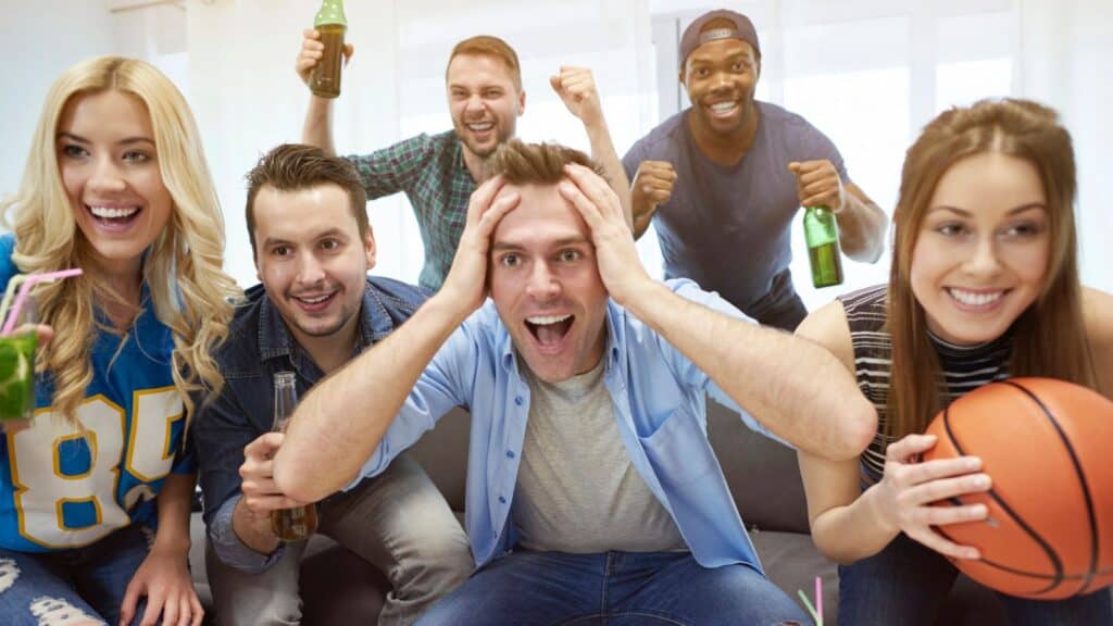 Five people sit on a couch, excitedly watching something off-camera. Some hold drinks; one person holds a basketball, and all appear engaged and enthusiastic.