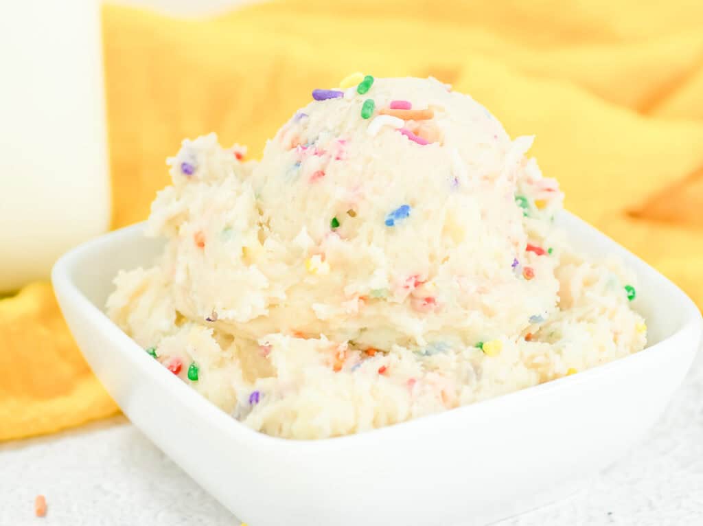A bowl of creamy funfetti cookie dough with colorful sprinkles, set against a yellow background.