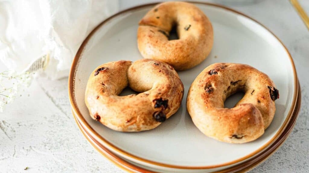 Three baked bagels with raisins are arranged on a white plate placed on a stack of plates.