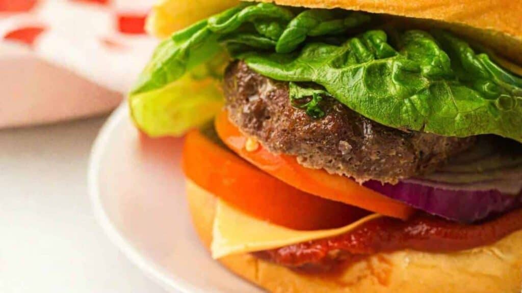 Close-up of a hamburger with lettuce, tomato, red onion, cheese, and ketchup on a bun, served on a white plate.