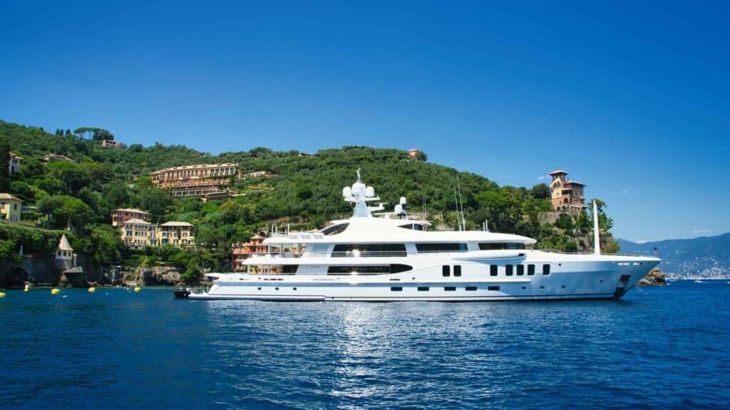 A large white yacht is anchored on calm blue water near a coastal area with green hills and villas in the background under a clear blue sky.