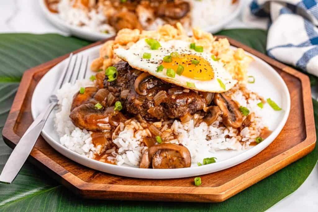 A plate of rice topped with hamburger patty, brown mushroom gravy, a fried egg, and chopped green onions, served with a fork on a wooden plate.