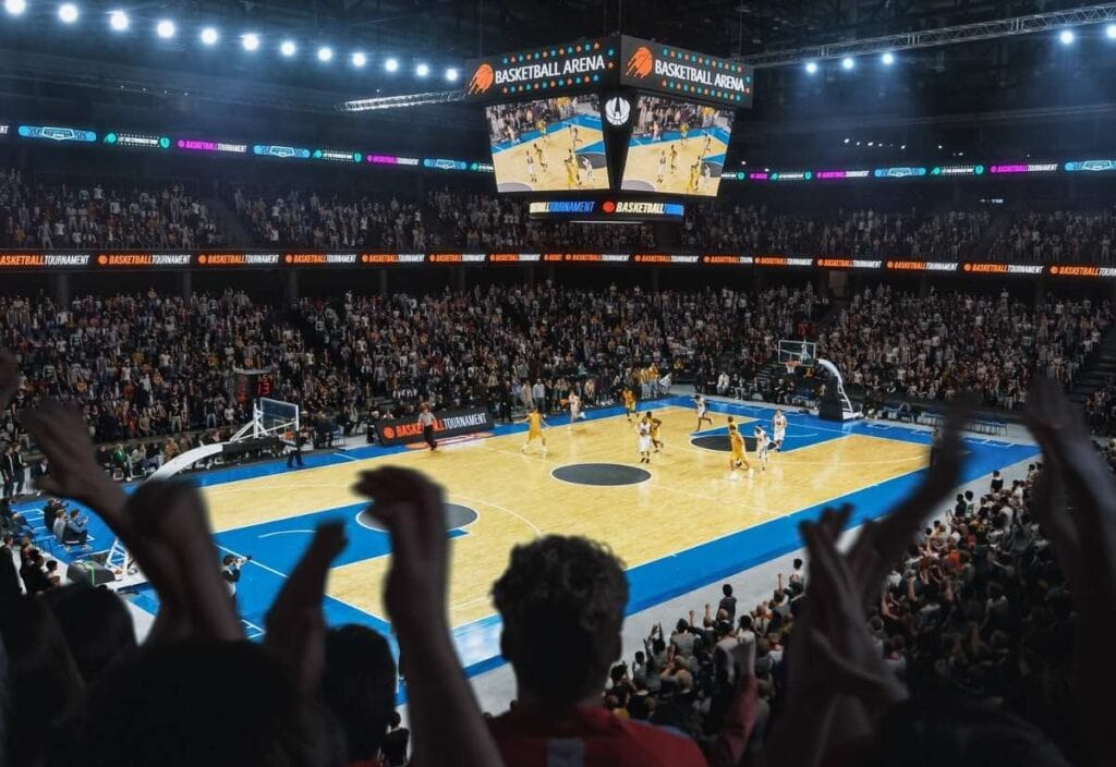 A packed indoor basketball arena buzzes with tournament travel excitement&mdash;a game in progress, large crowd cheering, and action displayed on overhead screens.