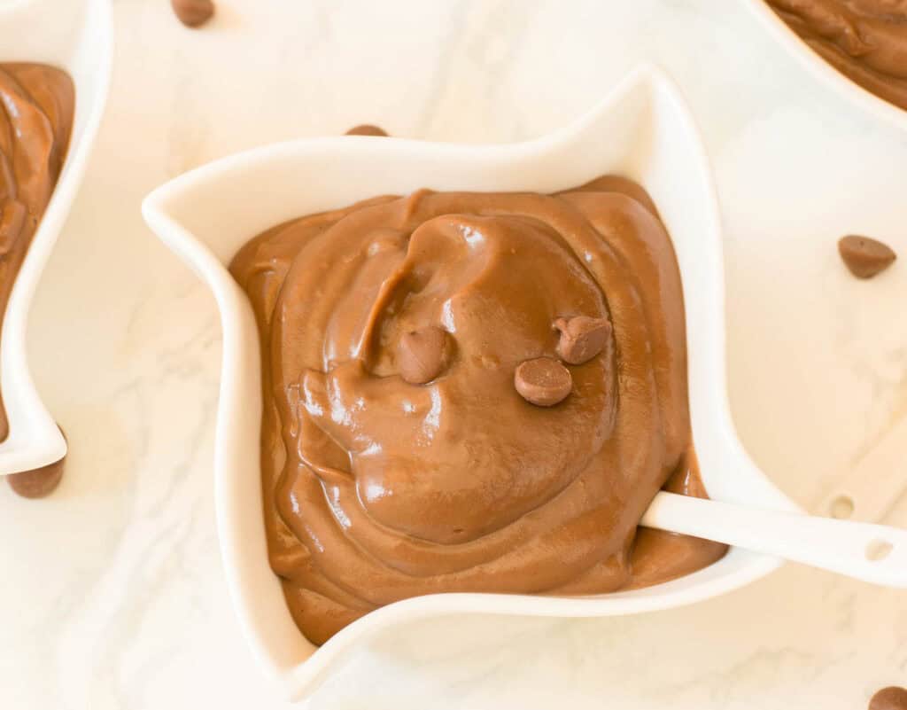 A white bowl filled with creamy chocolate pudding, topped with chocolate chips, with a white spoon resting inside the bowl.