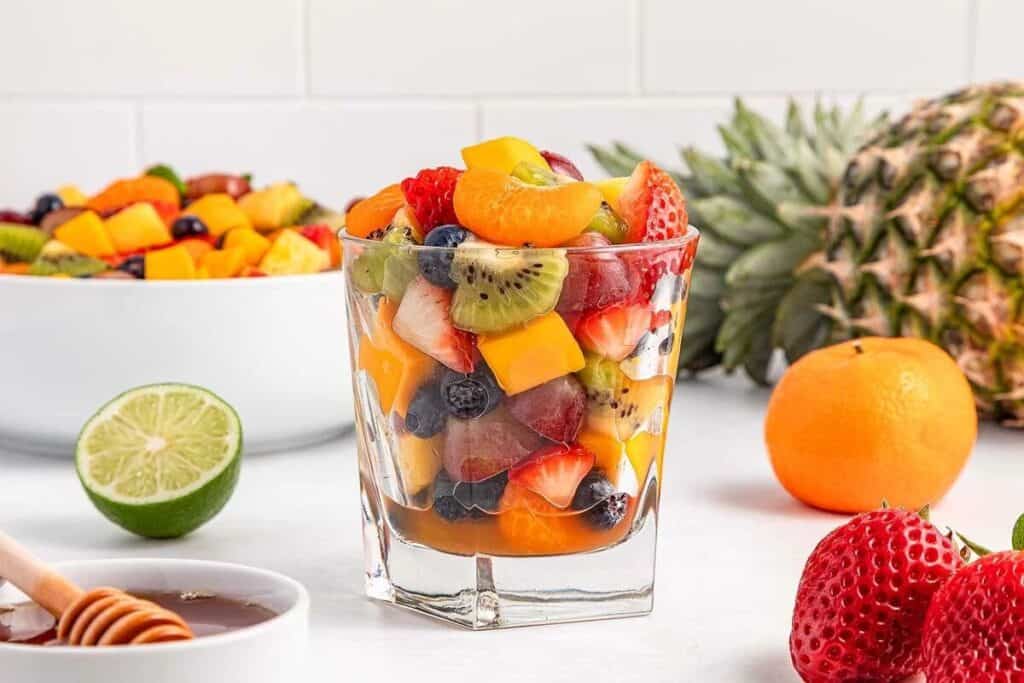 A glass filled with mixed fresh fruit pieces including strawberries, kiwi, grapes, and blueberries, with a bowl of fruit, honey, lime, orange, and pineapple in the background.