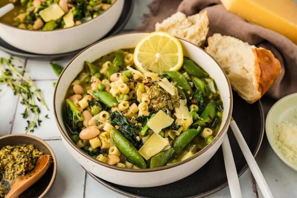 A bowl of vegetable soup with pasta, beans, snap peas, greens, pesto, and lemon slice, served with bread and garnished with cheese shavings.