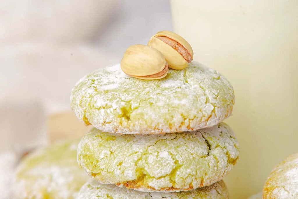 Three powdered sugar-covered green cookies are stacked, with two shelled pistachios placed on top.