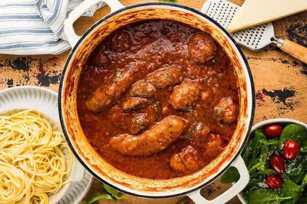 A pot of Italian sausage and meatballs in tomato sauce, next to a plate of cooked spaghetti and a bowl of salad with cherry tomatoes.