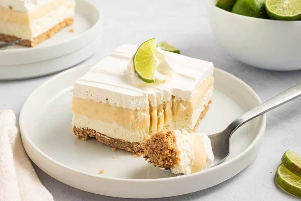 A slice of creamy layered citrus dessert with a graham cracker crust, topped with whipped cream and a lime wedge, is served on a white plate with a forkful taken out.