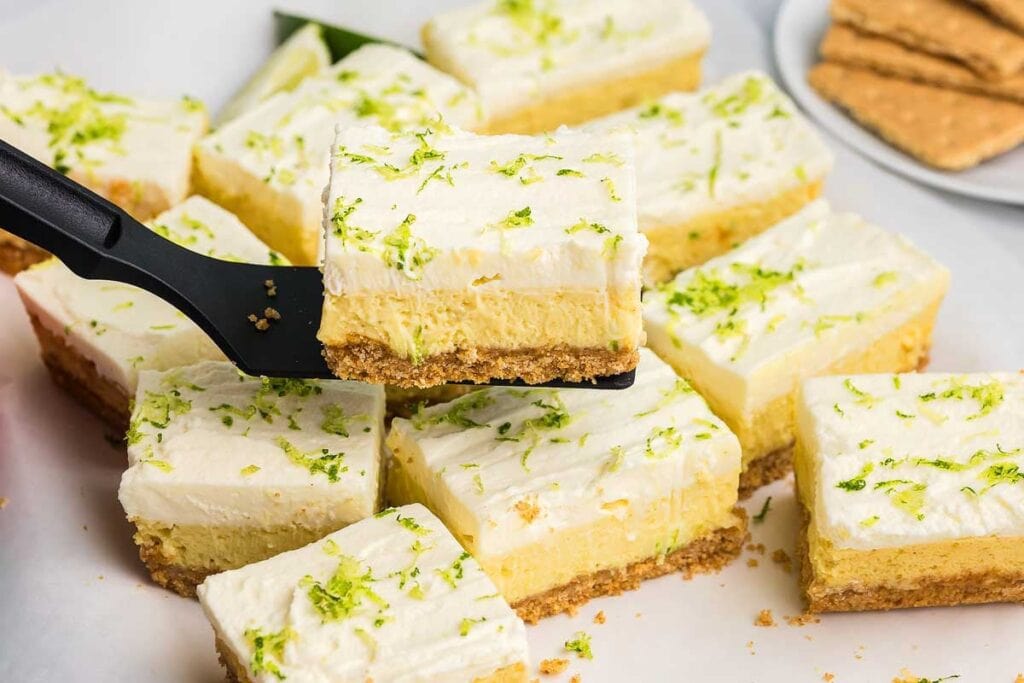 A spatula lifts a square piece of key lime pie bar with a graham cracker crust, creamy filling, whipped topping, and lime zest. Several bars are arranged on parchment paper.