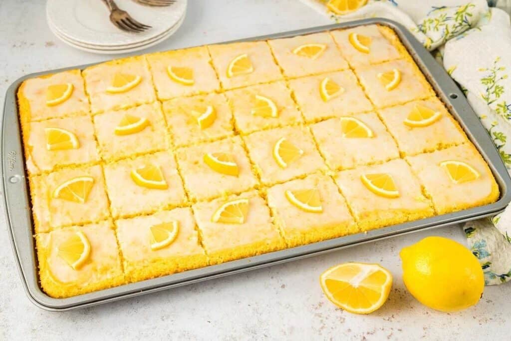 A rectangular pan of lemon sheet cake, cut into squares, each topped with a small lemon wedge. A whole lemon and lemon wedges are beside the pan. Plates and forks are in the background.