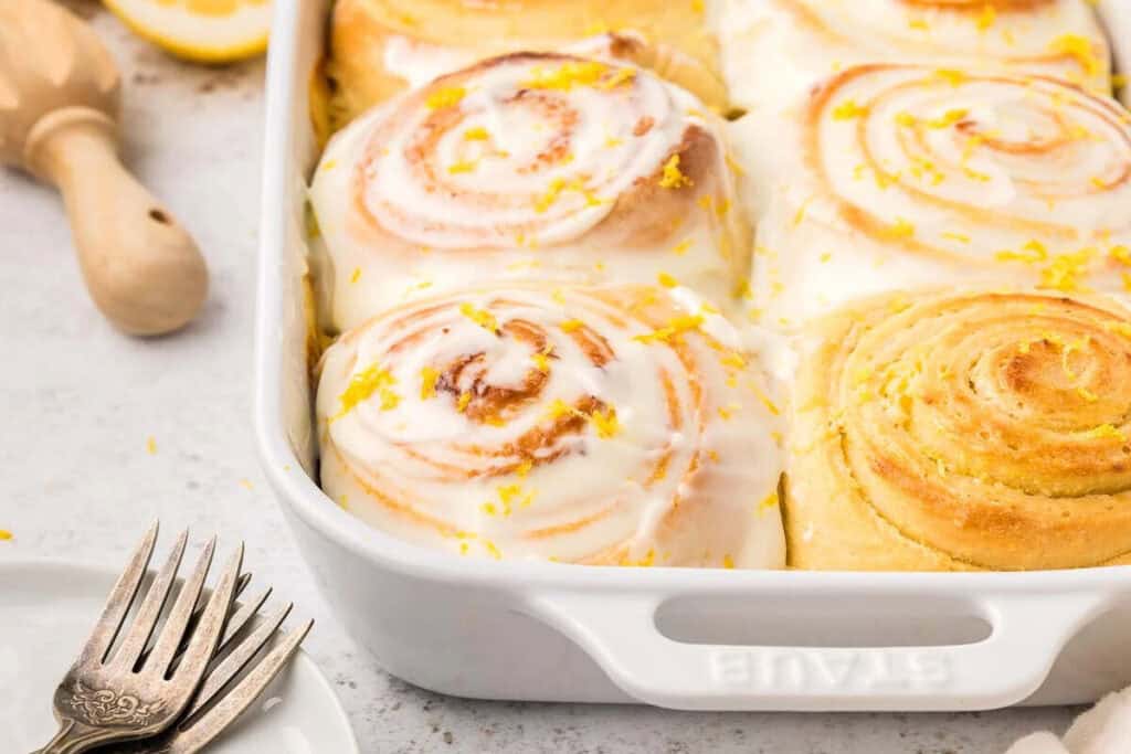 Iced cinnamon rolls topped with lemon zest in a white baking dish, with three forks and a citrus reamer nearby on a light countertop.