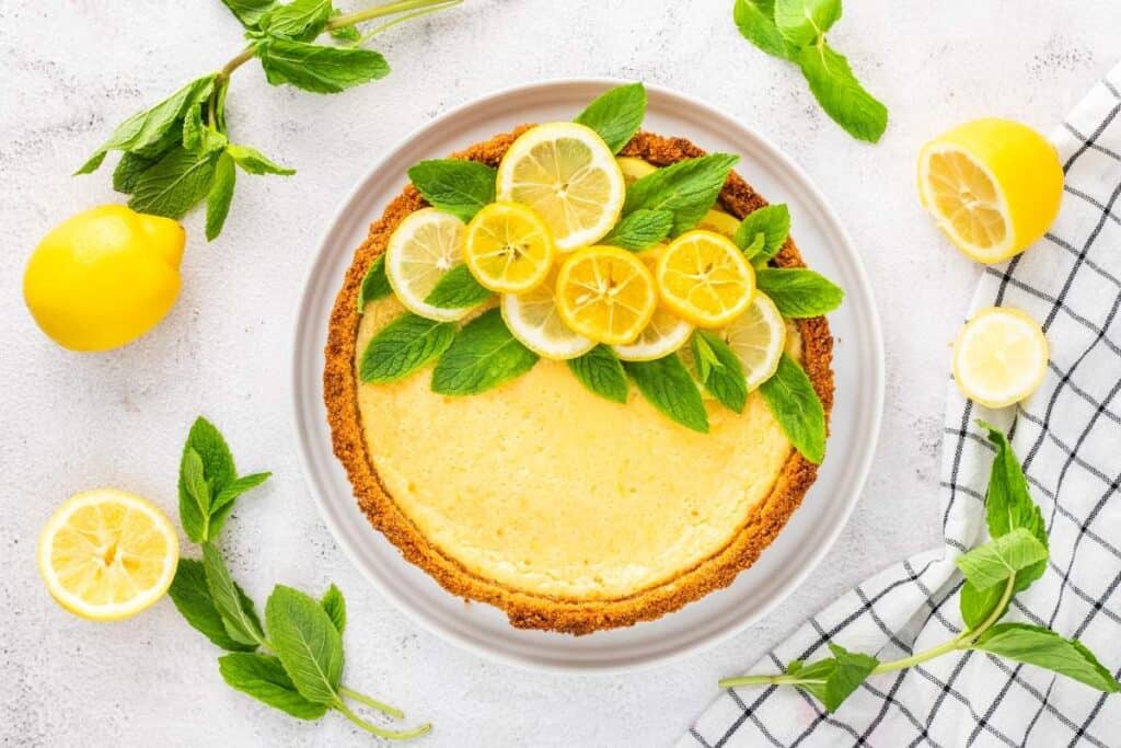A lemon tart, a classic among citrus desserts, is topped with fresh mint leaves and lemon slices on a white plate, surrounded by lemons and mint on a light surface.