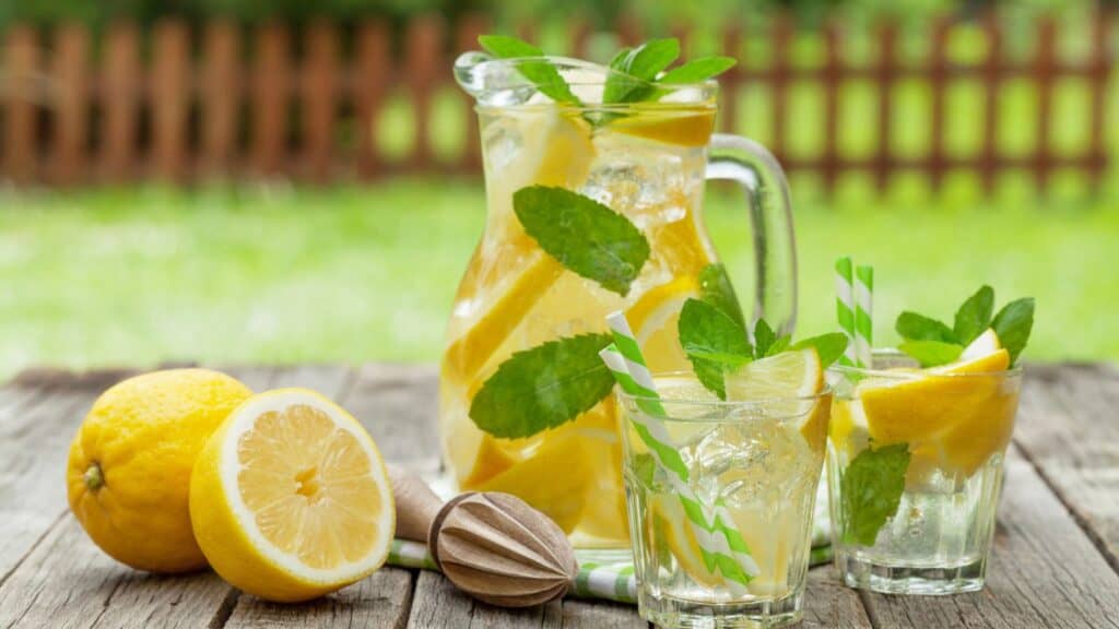 A pitcher and two glasses of lemonade with lemon slices and mint leaves sit on a wooden table outdoors, next to whole and halved lemons and a citrus reamer.