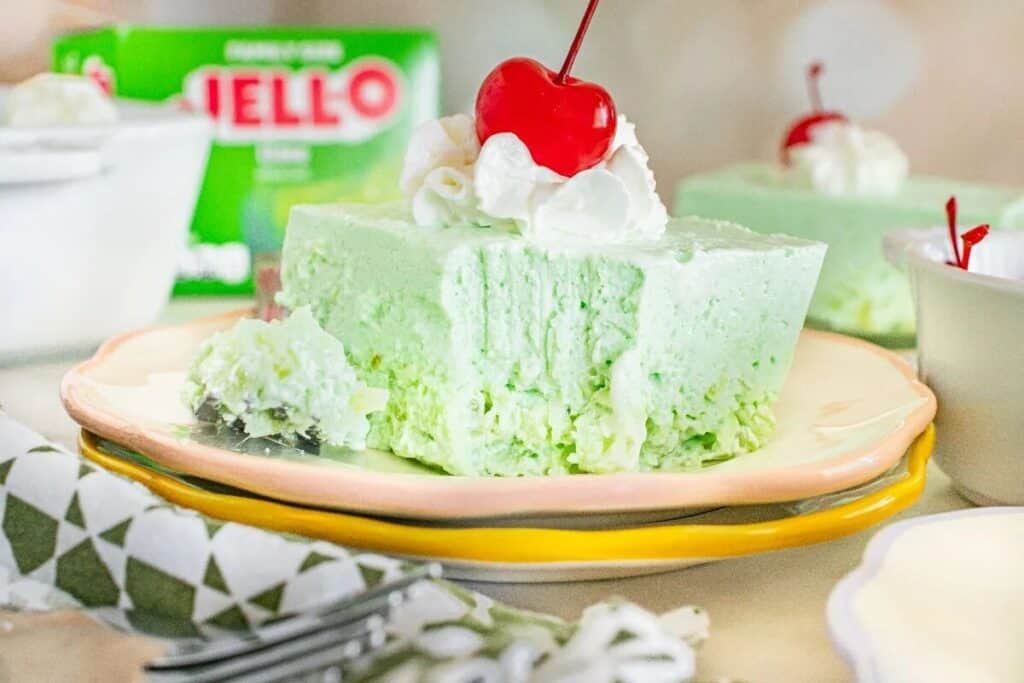 A slice of light green Jello citrus dessert topped with whipped cream and a cherry sits on a plate, with a forkful taken out. A box of lime Jello is visible in the background.