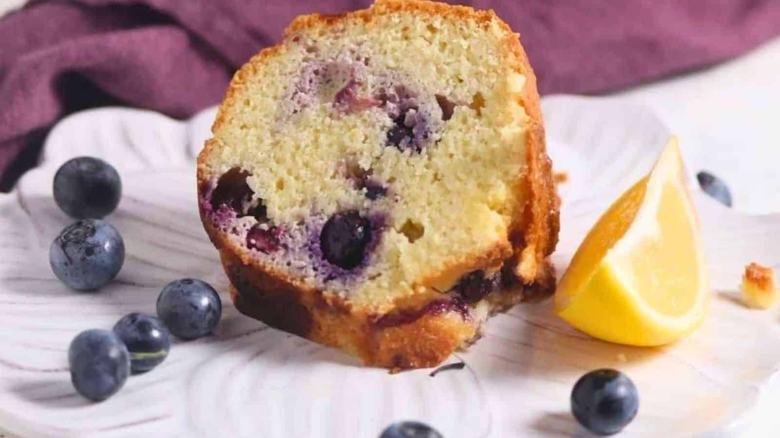 A slice of blueberry bundt cake sits on a plate with fresh blueberries and a lemon wedge beside it.