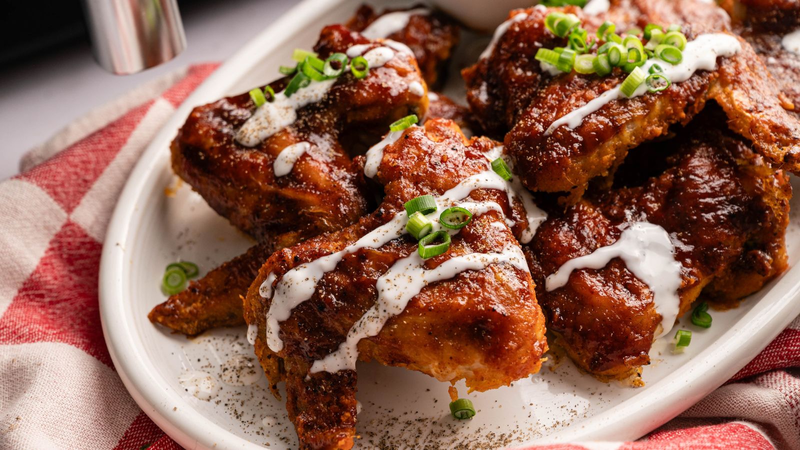 A plate of saucy chicken wings garnished with chopped green onions and a drizzle of white sauce, served on a white dish with a red and white napkin underneath.