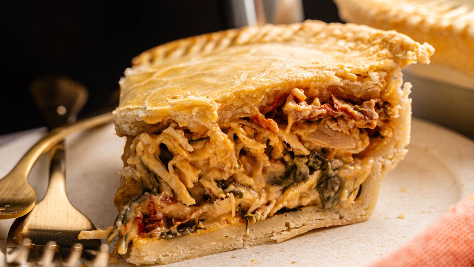 A close-up of a slice of savory pie filled with creamy shredded chicken, vegetables, and a golden flaky crust, served on a white plate with a fork and knife.