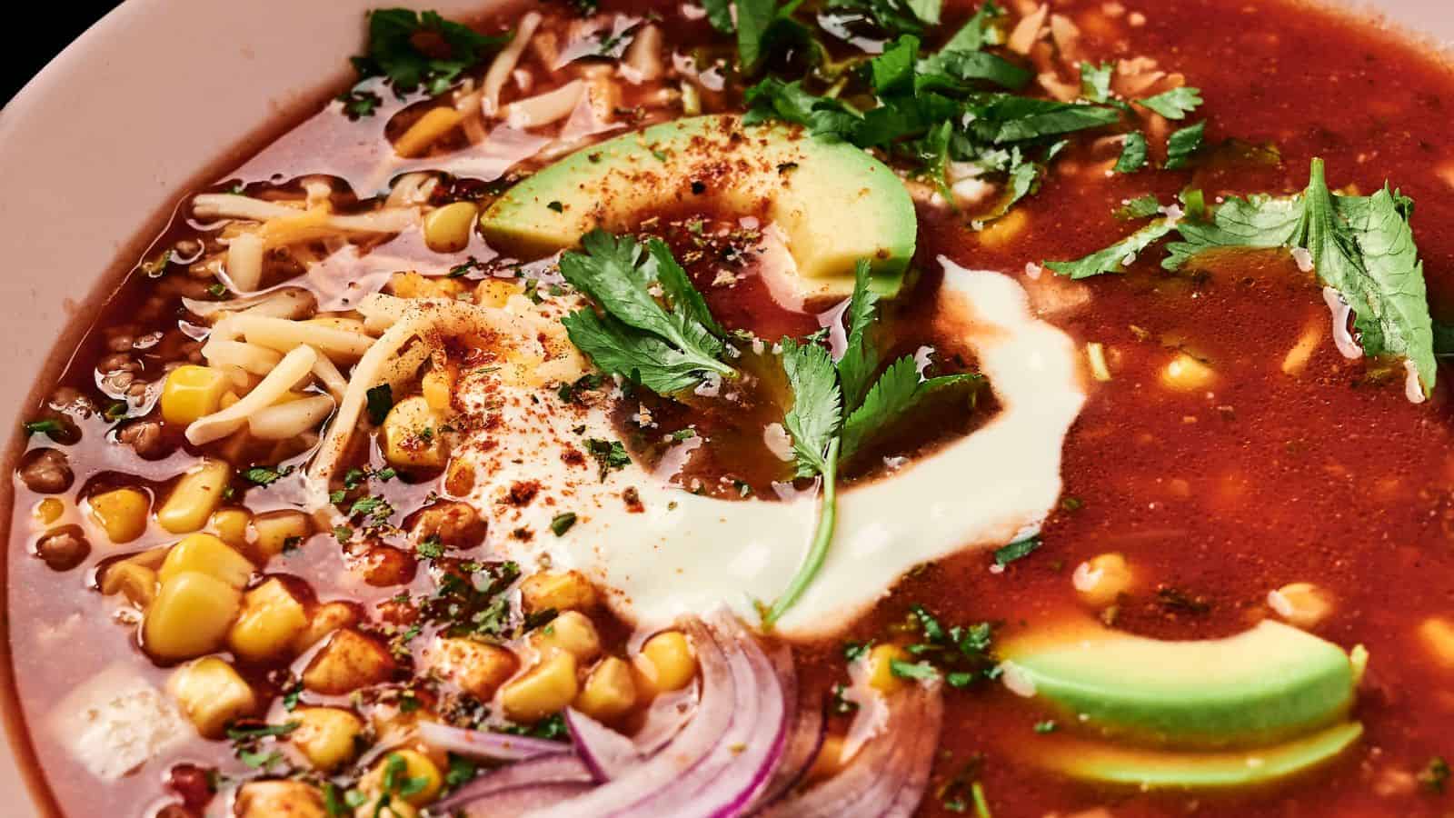 Close-up of a bowl of tortilla soup topped with sliced avocado, corn, shredded cheese, red onion, cilantro, and a dollop of sour cream.