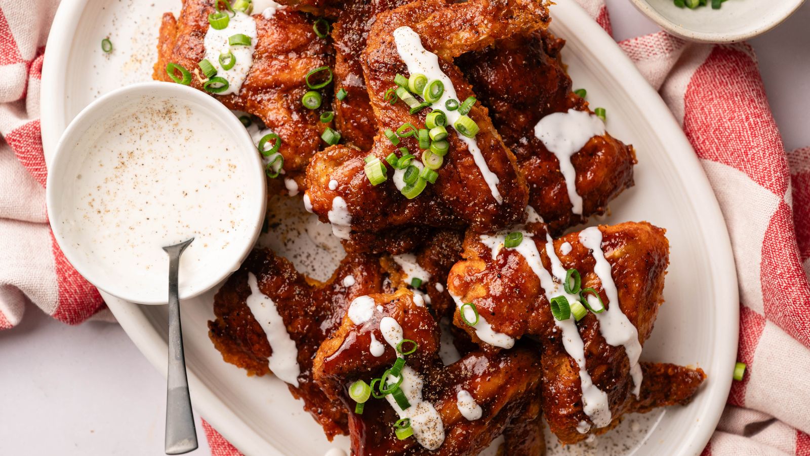 A platter of glazed chicken wings topped with chopped green onions and drizzled with white sauce, served with a small bowl of dipping sauce and a spoon.