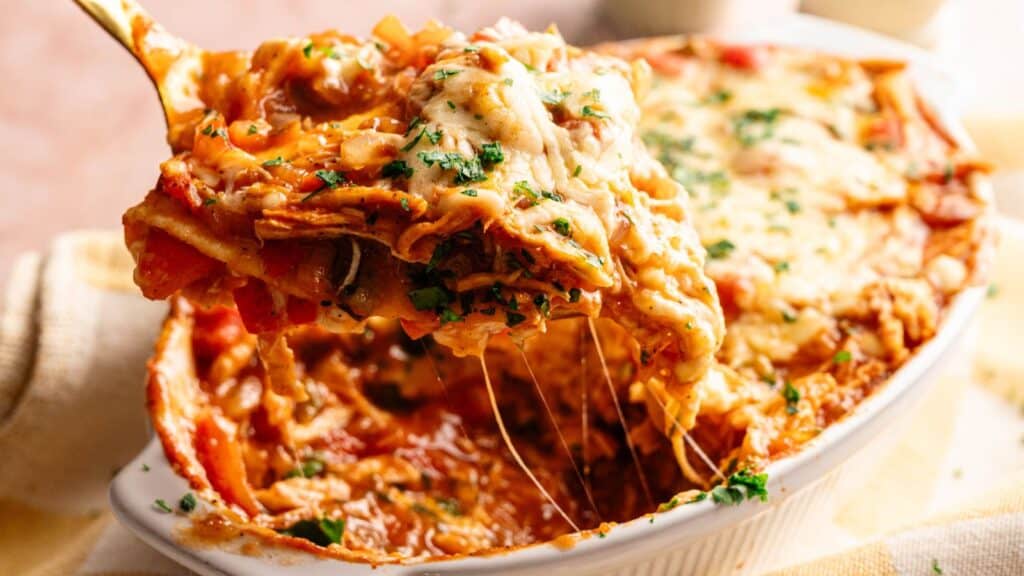 A serving of cheesy lasagna is being lifted from a baking dish, showing layers of pasta, sauce, vegetables, and melted cheese, topped with chopped herbs.