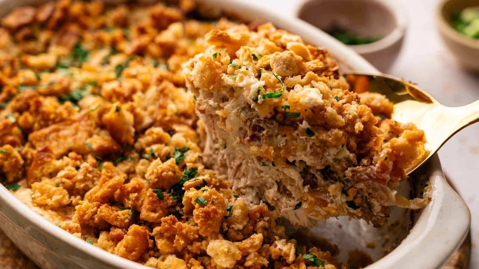 A serving spoon lifts a portion of a baked casserole topped with golden, crispy breadcrumbs and garnished with chopped herbs from a white dish.