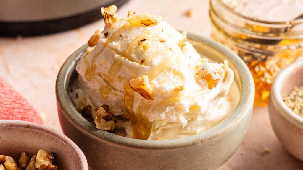 A bowl of vanilla ice cream topped with chopped nuts and drizzled with honey, with jars of honey and nuts in the background.