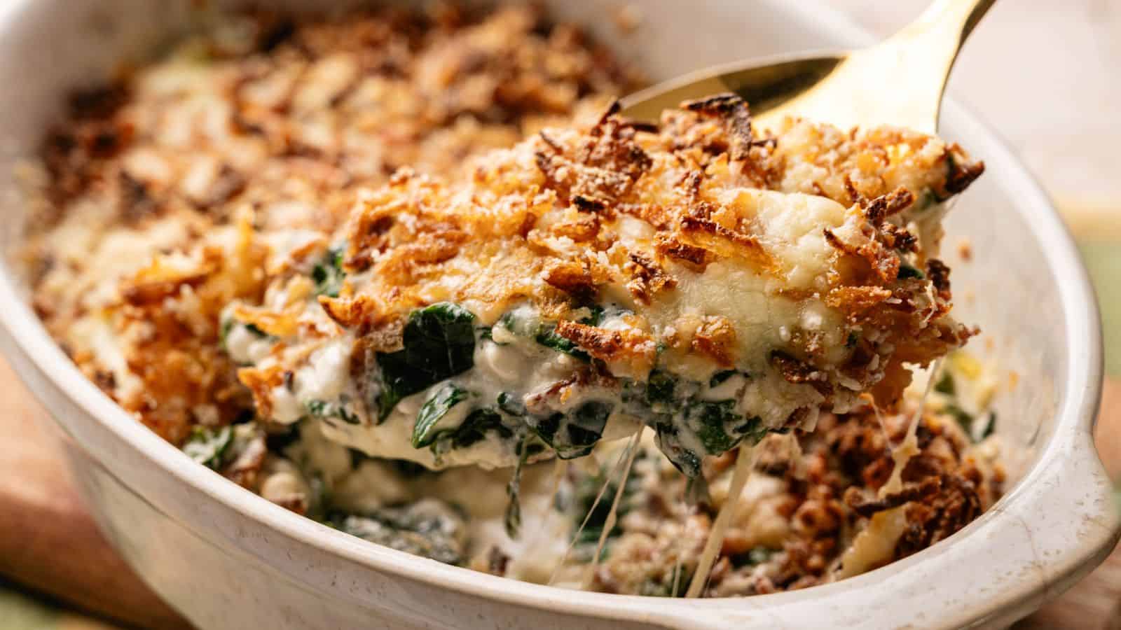 A serving of creamy baked spinach casserole with a crispy breadcrumb topping is being lifted from a white baking dish with a gold spoon.